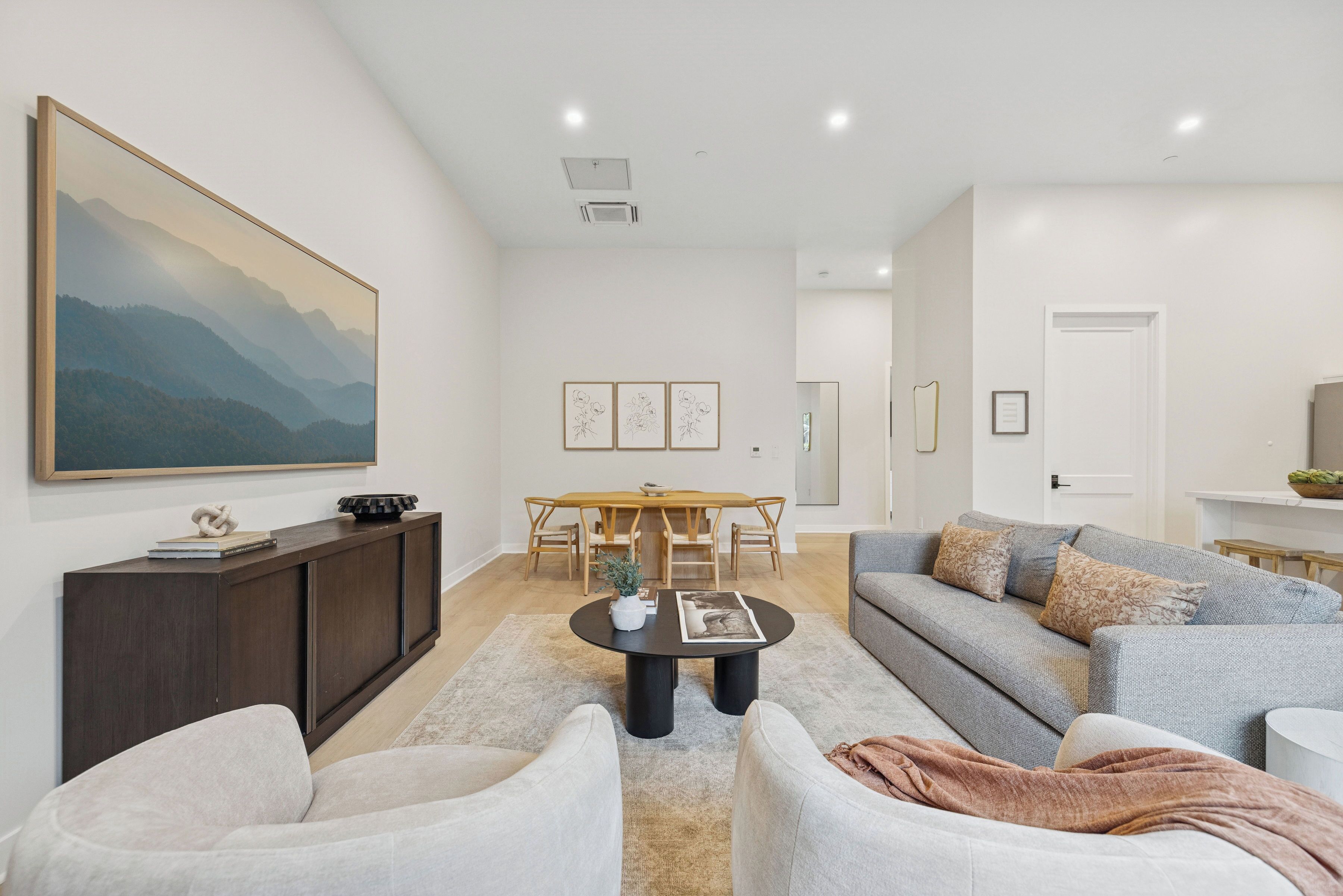 A modern living room with a large painting, a dining area with a wooden table and chairs, and a grey sofa with decorative pillows.