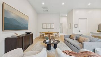 A modern living room with a large painting, a dining area with a wooden table and chairs, and a grey sofa with decorative pillows.