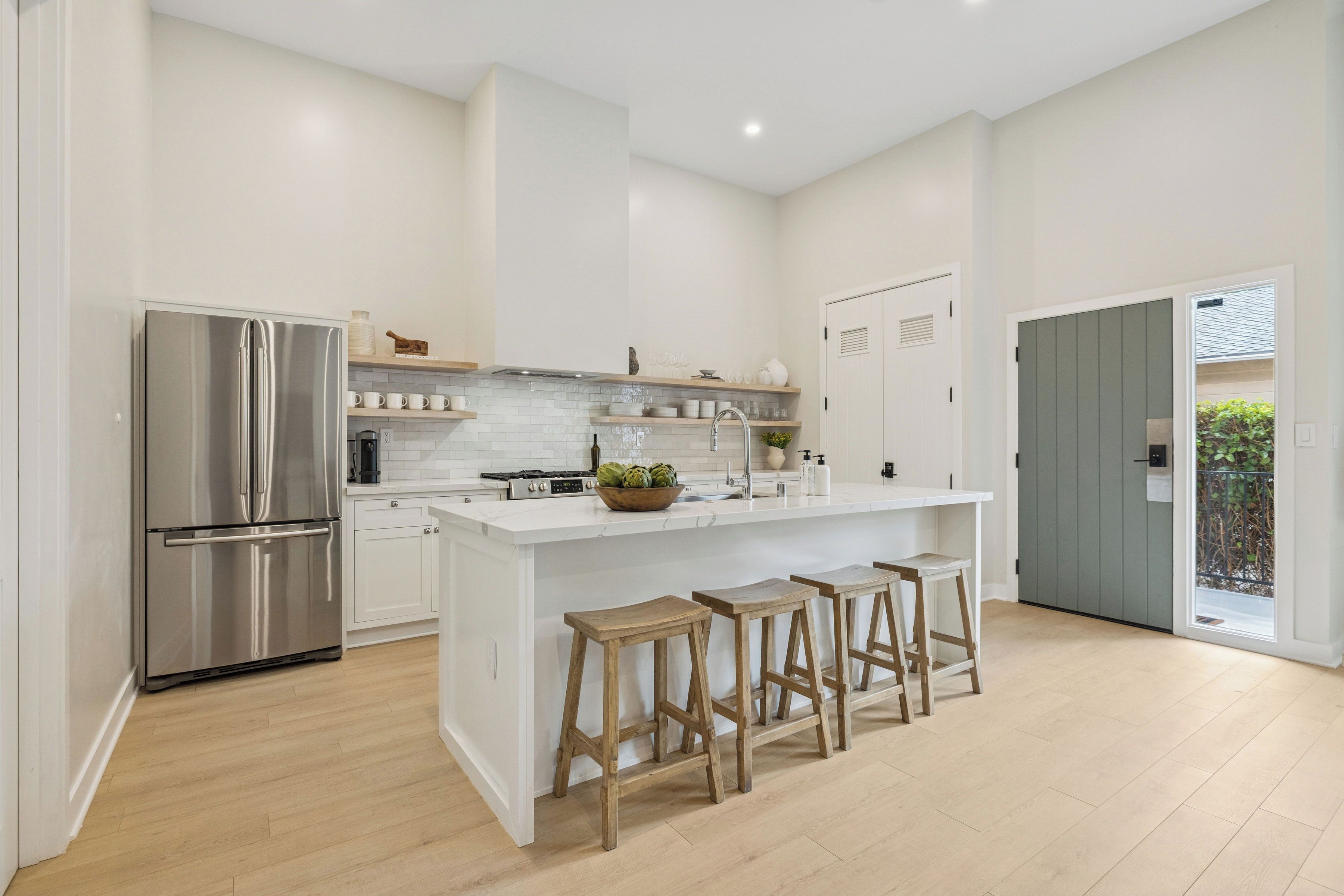 A modern kitchen with a stainless steel refrigerator, white cabinetry, a central island with stools, and a view of the outdoors through a door.