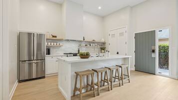A modern kitchen with a stainless steel refrigerator, white cabinetry, a central island with stools, and a view of the outdoors through a door.