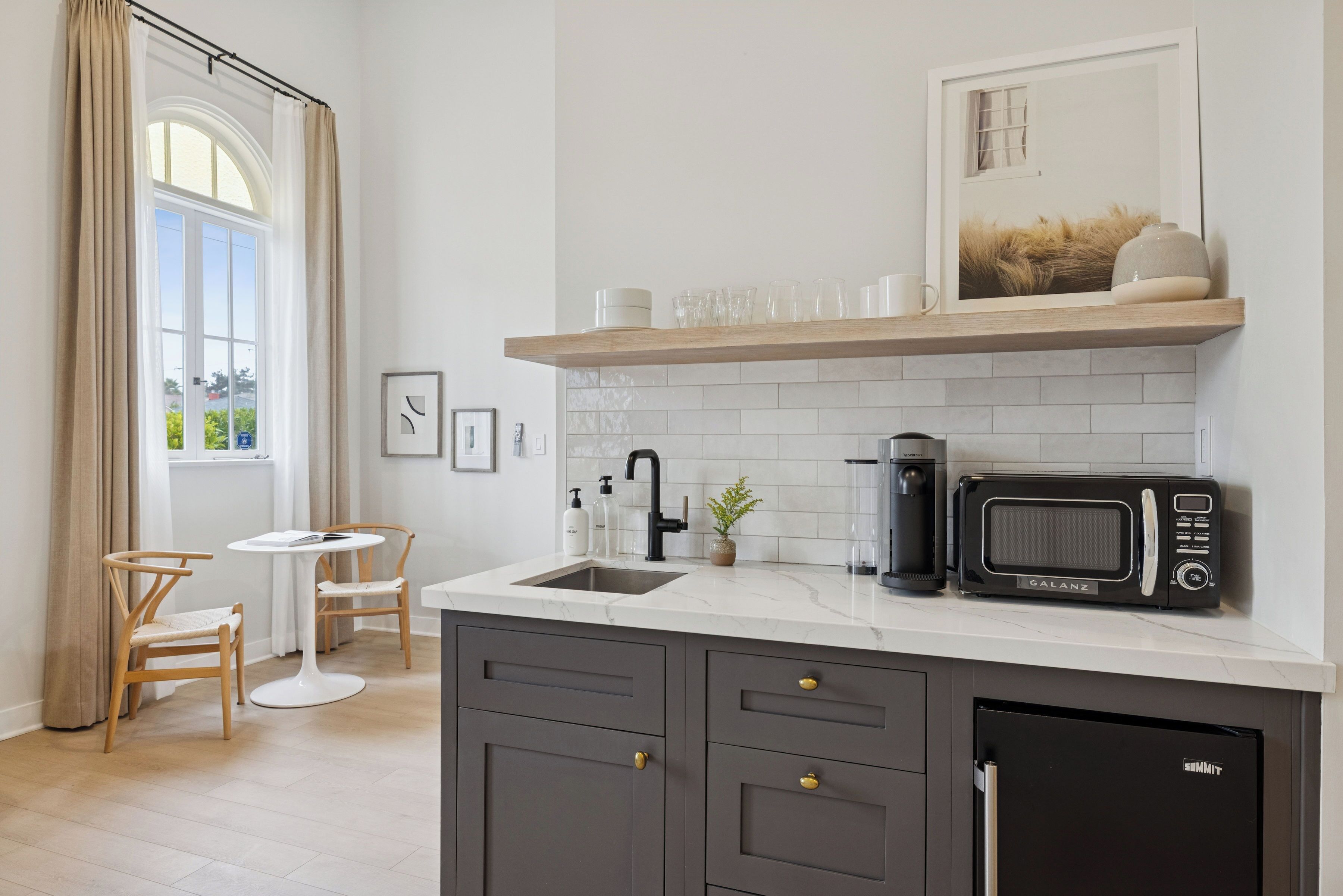 Cocina compacta con lavamanos, cafetera, microondas y refrigerador. Un pequeño comedor con dos sillas y una mesa.