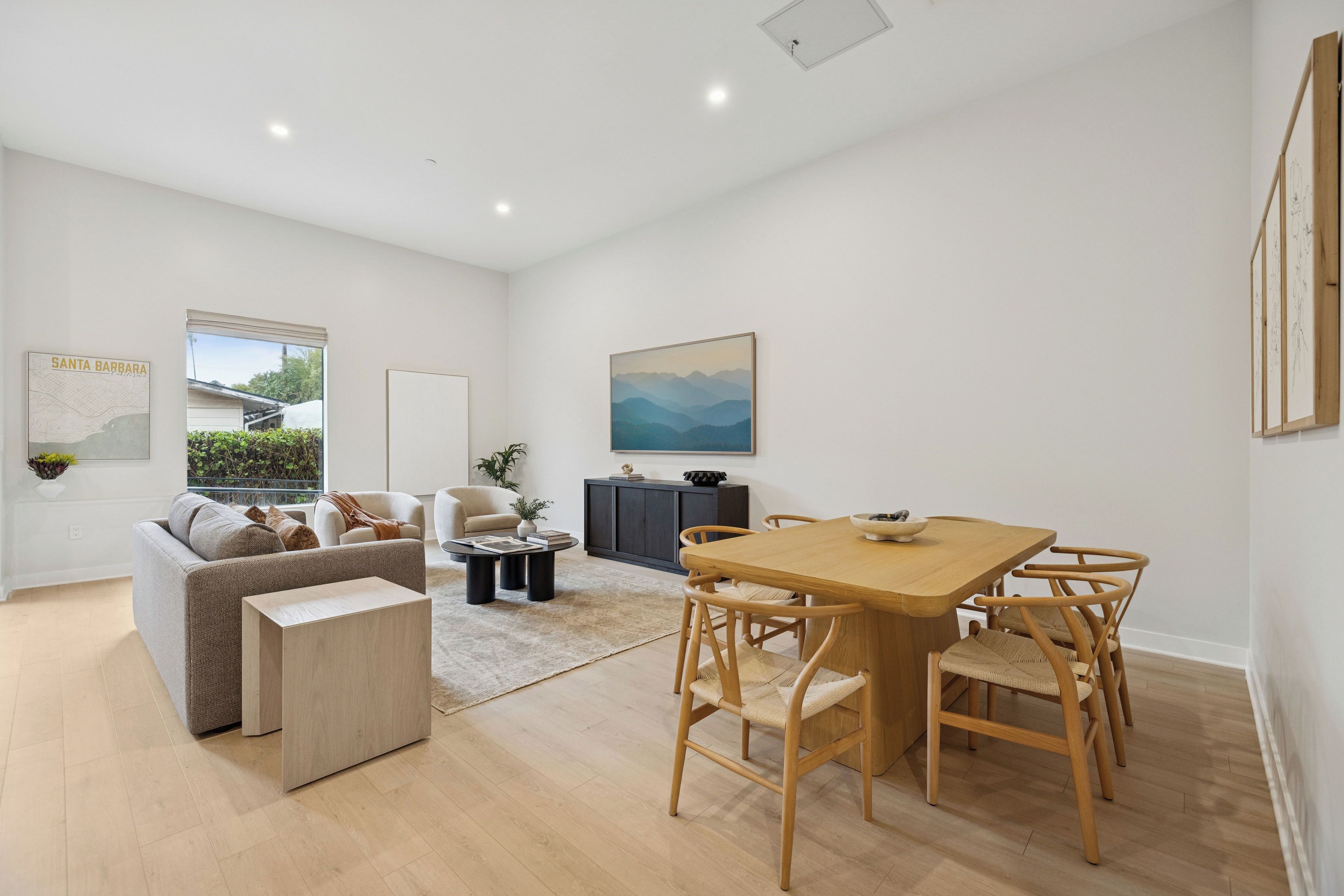 A modern living room with a sofa, coffee table, and dining table with chairs. There is a window with a view of greenery and a painting on the wall.