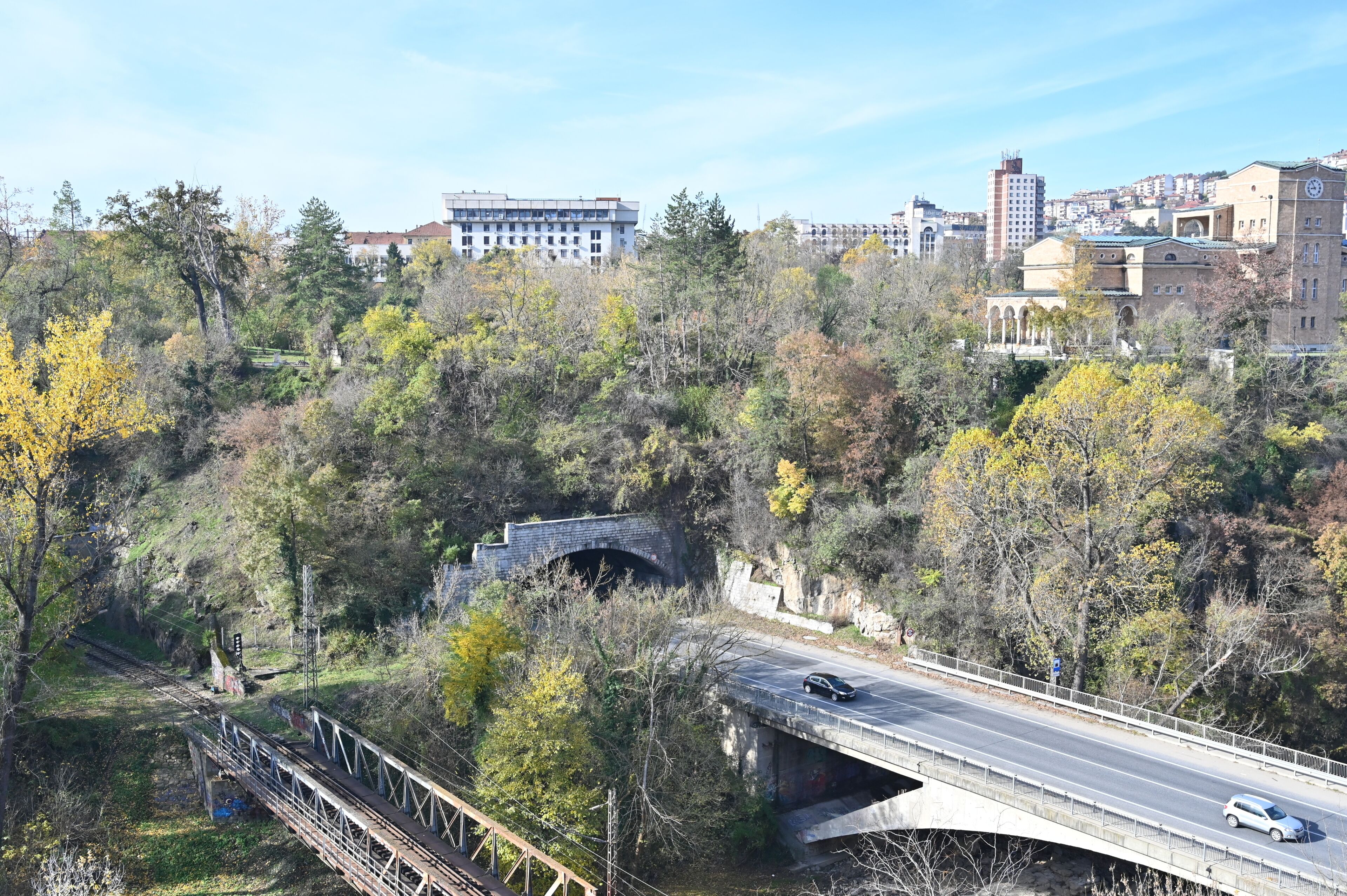 A bridge over a river with a road passing underneath, surrounded by trees and buildings in the background.