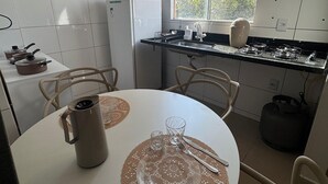 A modern kitchen with white tiles, a round dining table with a coffee pot and cutlery, and a window with a view of buildings and trees. - Residencial Gomes (Goiânia)