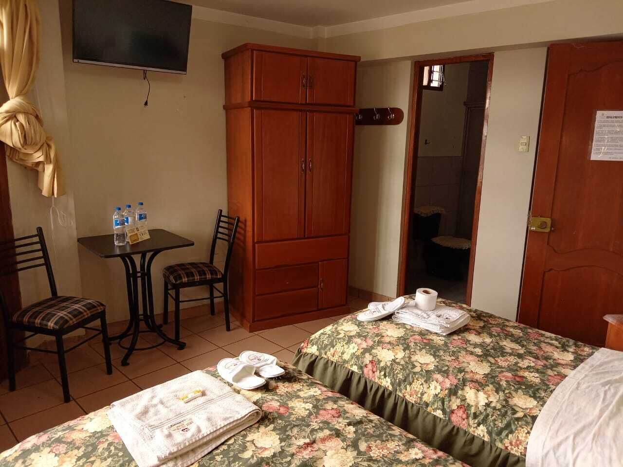 Habitació d'hotel amb dos llits, un armari de fusta, una taula petita amb cadires, un televisor fixat a la paret i una porta que condueix a una altra habitació.