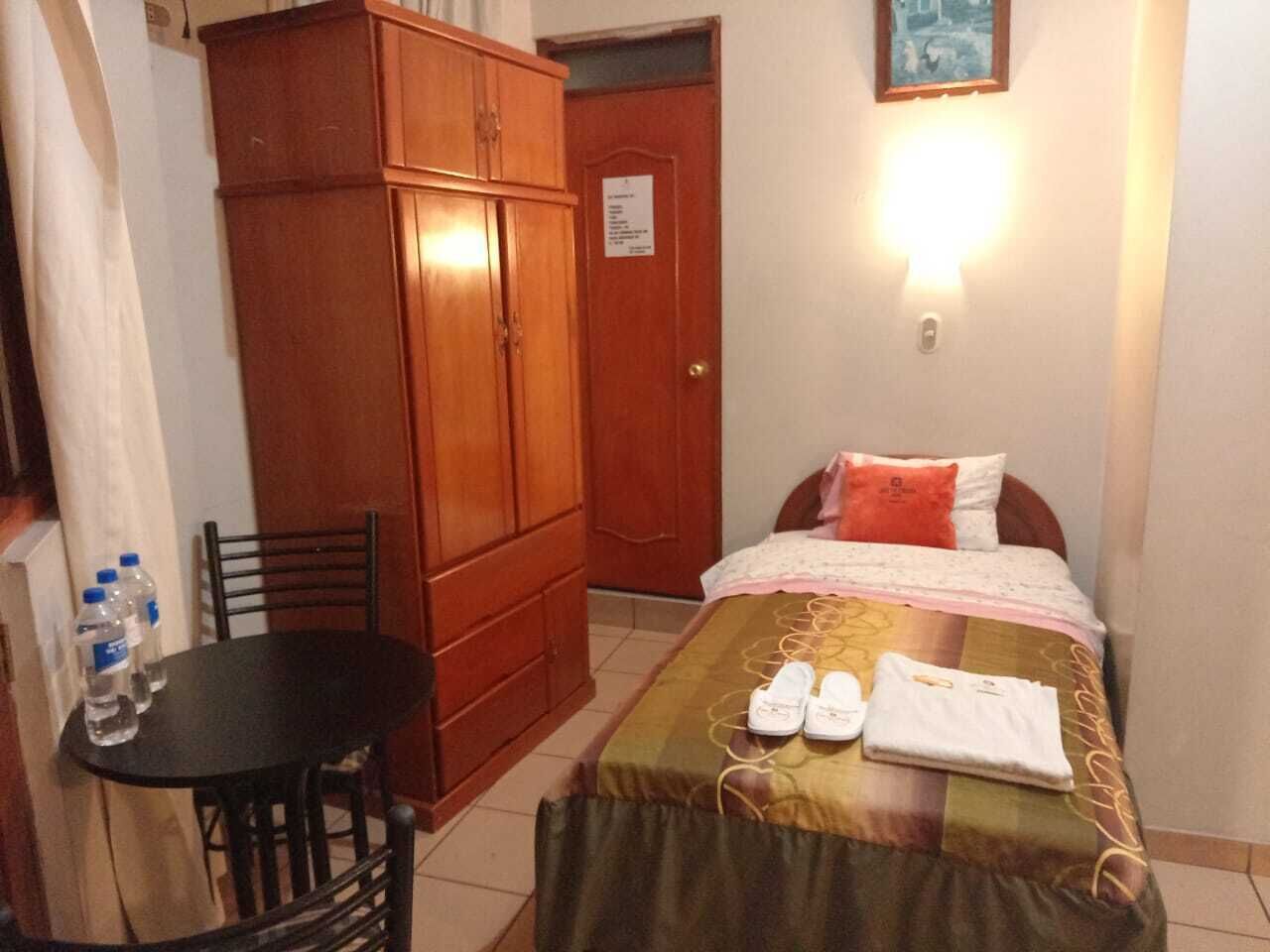 A bedroom with a bed, a wooden wardrobe, a table with water bottles, and a wall-mounted light.