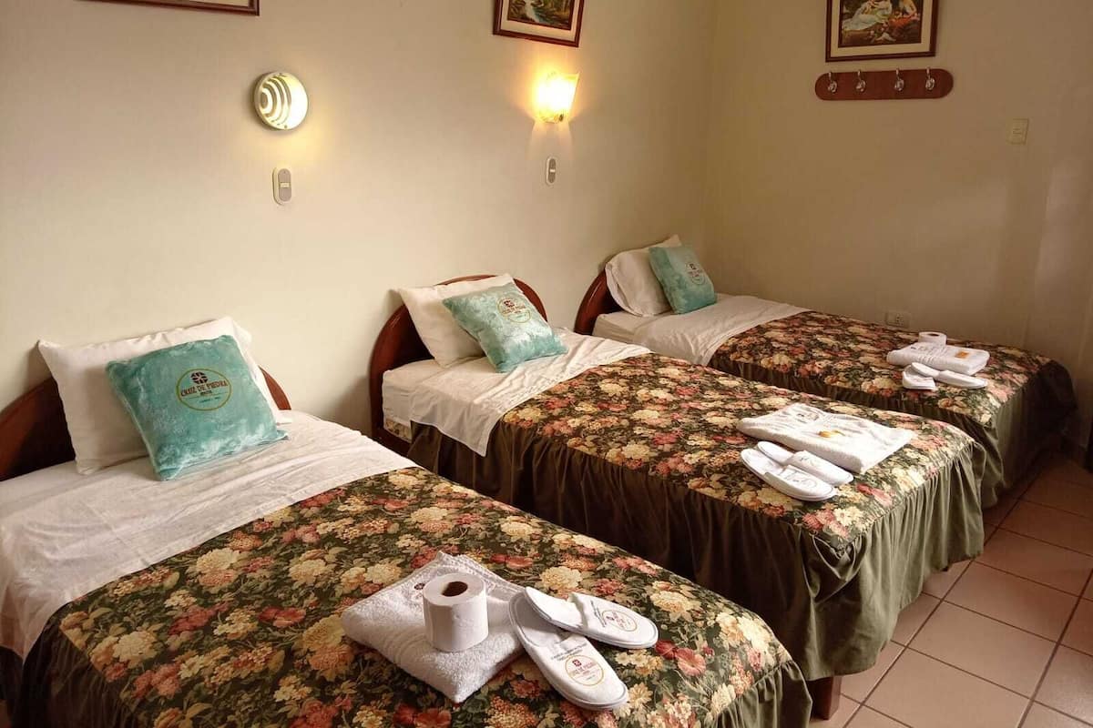 Une chambre d’hôtel avec trois lits simples, chacun agrémenté d’une couvre-lit à motifs floraux et de serviettes. Les murs sont ornés d’appliques murales et de tableaux encadrés.