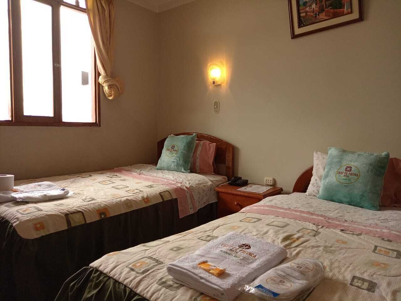 A hotel room with two beds, a bedside table with a lamp, a window with curtains, and a wall-mounted light.