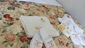 A bed with a floral bedspread, two red pillows, a white towel, and a pair of slippers with a logo.