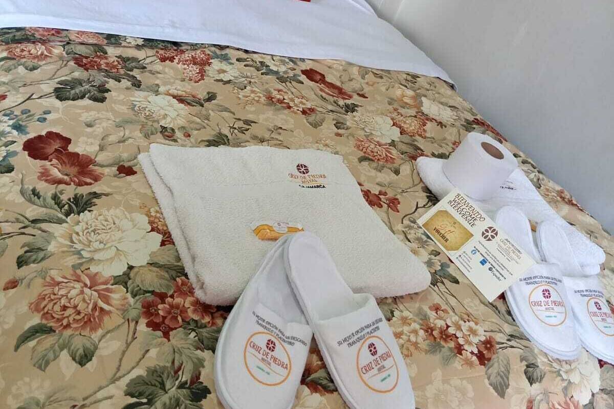 Un lit avec une couvre-lit à motifs floraux, deux oreillers rouges, une serviette blanche et une paire de chaussons ornés d’un logo.