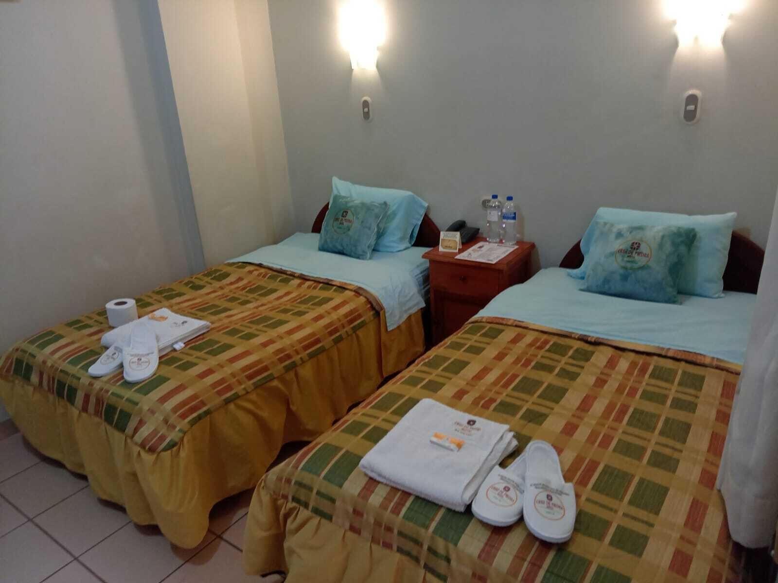 A hotel room with two beds, each with a checkered bedspread, pillows, and towels. There is a nightstand with a lamp and a phone between the beds.