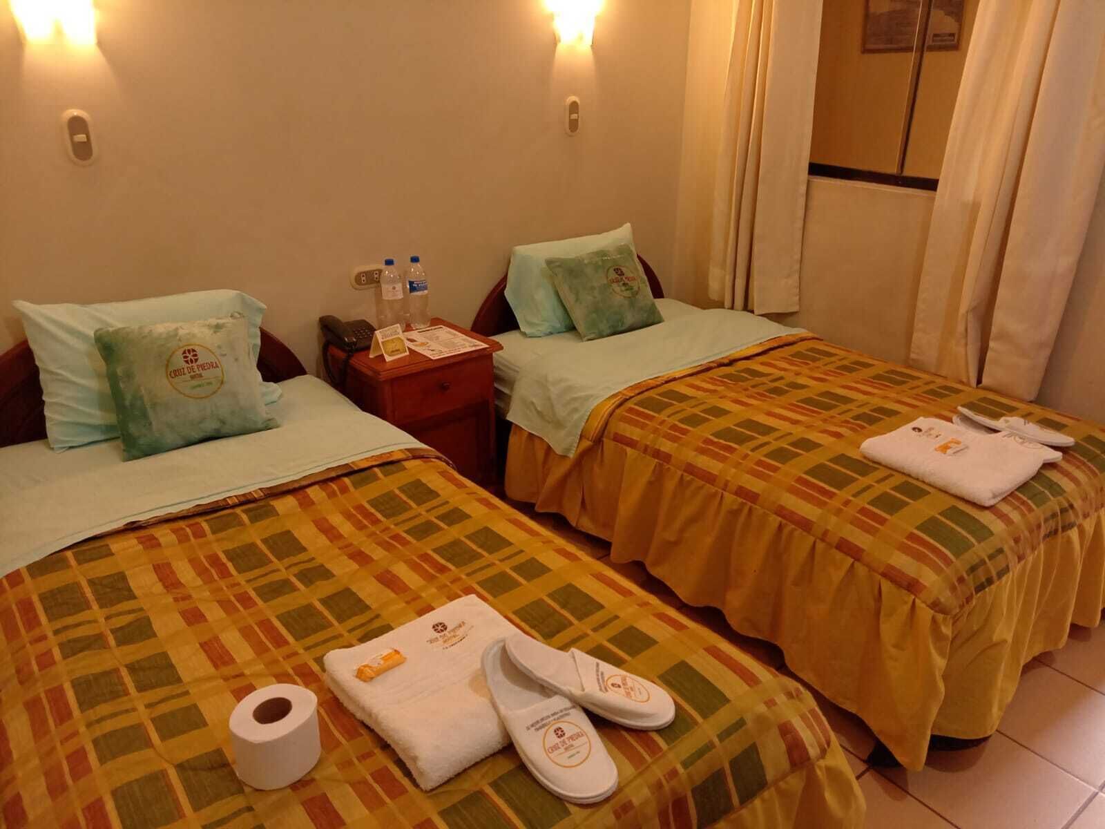 Habitación de hotel con dos camas, cada una con almohada y manta. Hay artículos de tocador en la mesita de noche, un teléfono y una ventana con cortinas.