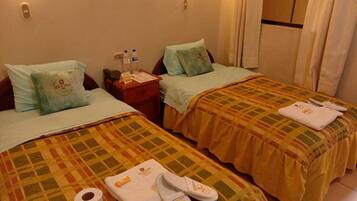 Habitación de hotel con dos camas, cada una con almohada y manta. Hay artículos de tocador en la mesita de noche, un teléfono y una ventana con cortinas.