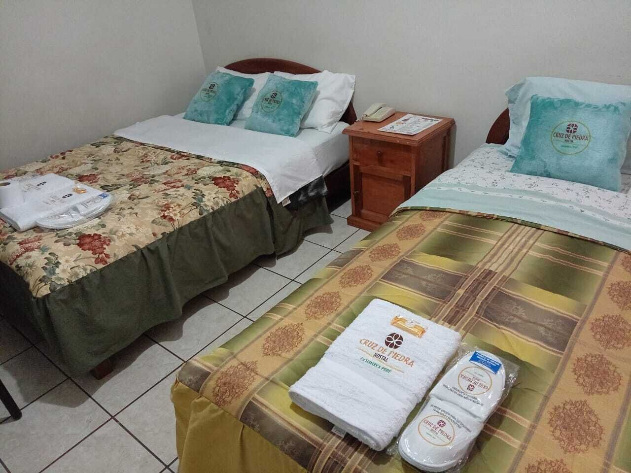 Camera d'albergo con due letti, ognuno con biancheria e cuscini floreali. Un comodino in legno con un telefono e un piccolo tavolo con un menu o un foglio informativo. Asciugamano e flacone di lozione sono posizionati sul letto.