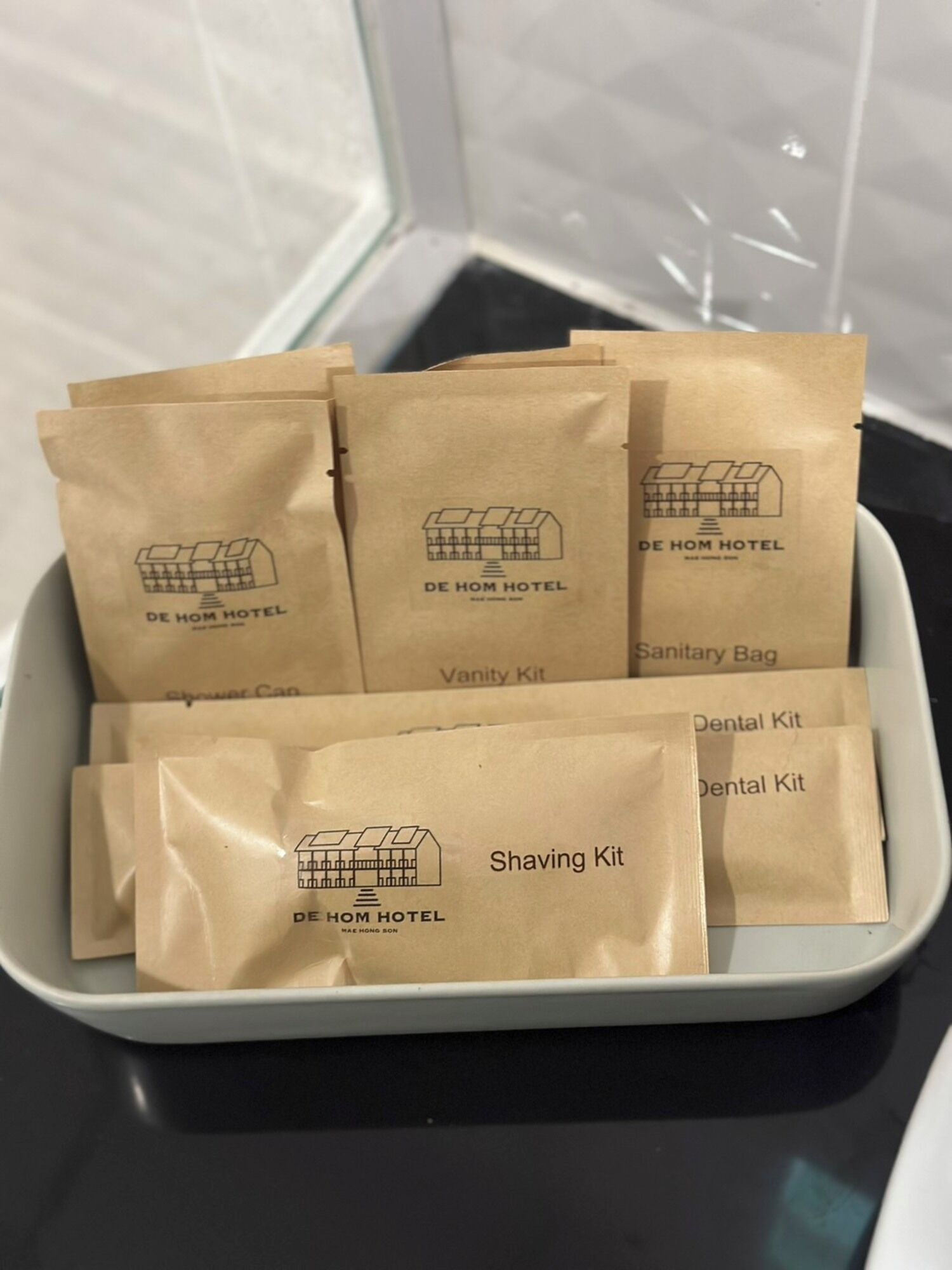 Een dienblad met verschillende bruine papieren zakken, met het logo van De Hom Hotel. De zakken zijn gelabeld als 'Sanitary Bag', 'Vanity Kit', 'Dental Kit' en 'Shaving Kit'.