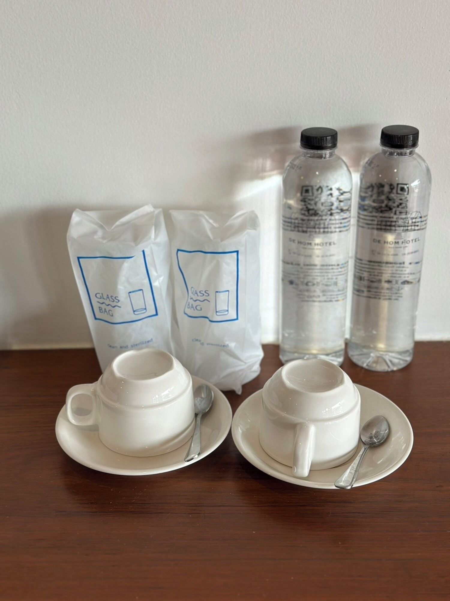 A table with two white cups and saucers, two glass bags labeled "GLASS BAG," and two bottles of water.