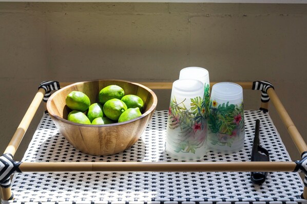 A wooden tray with a bowl of limes and two decorative glass containers on a woven table.