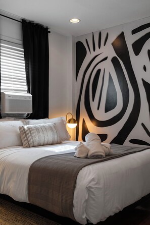 A hotel room with a bed, a wall-mounted air conditioner, a wall art piece, and a bedside lamp.