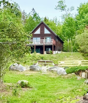 A wooden cabin with a balcony, surrounded by greenery and rocks.