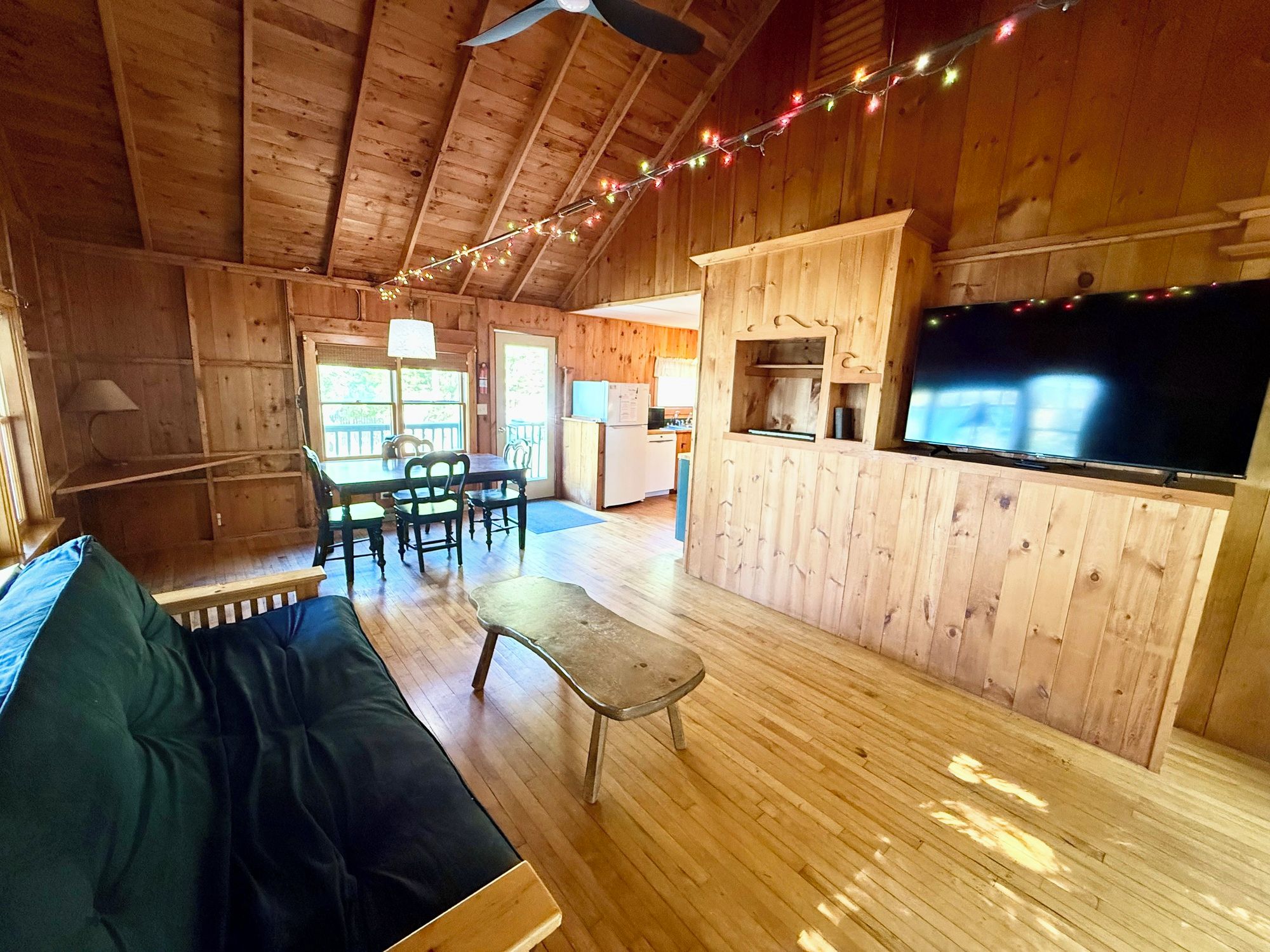 A wooden cabin interior with a sofa, dining table, chairs, a kitchen area, and a television.