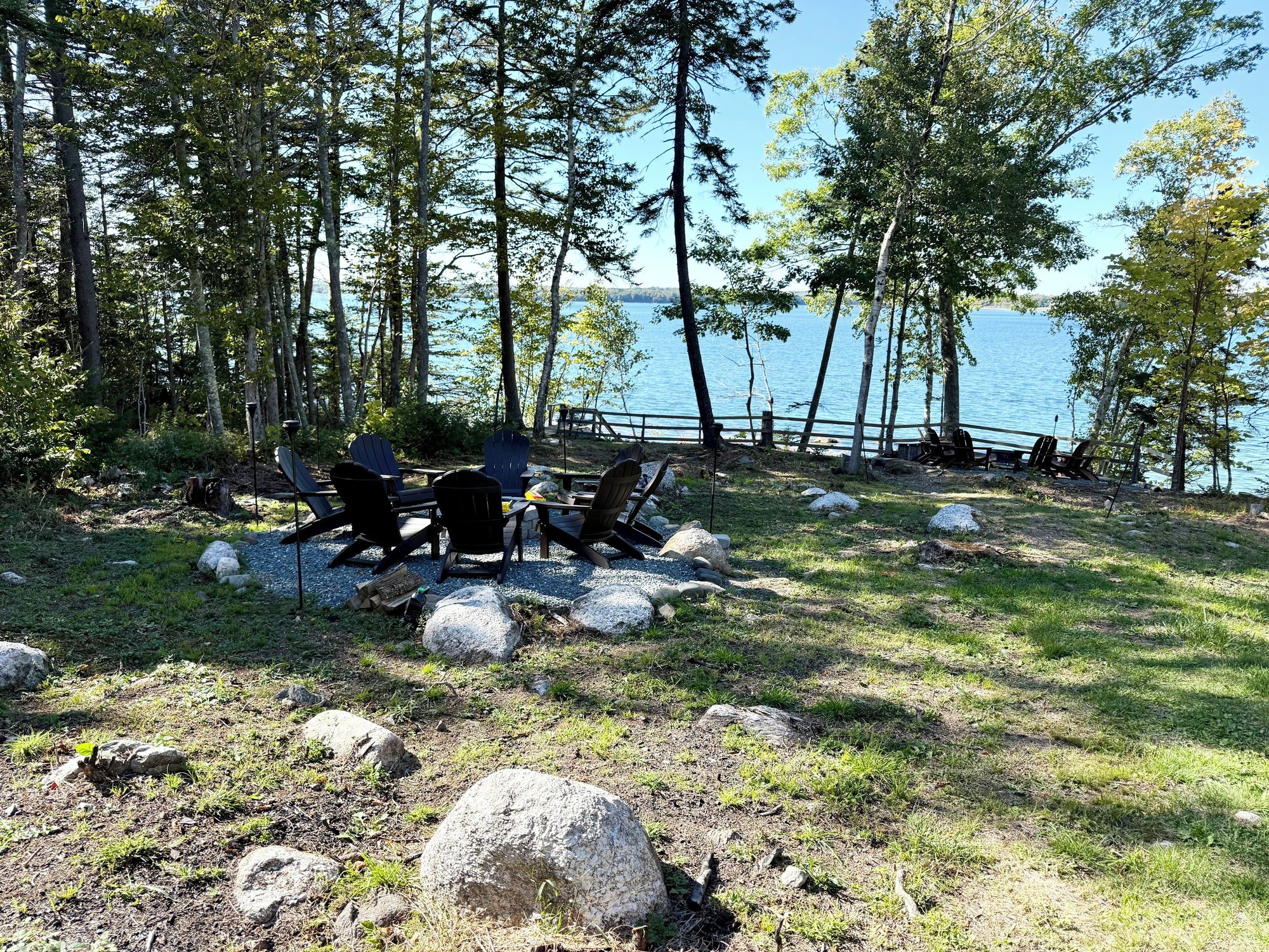 A lakeside camping area with Adirondack chairs and a fire pit.