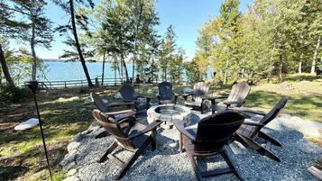 An outdoor seating area with Adirondack chairs and a fire pit, surrounded by trees and a lake view.