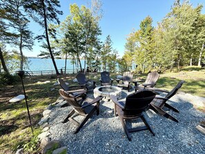 Et udendørs opholdsområde med Adirondack-stole og bålsted, omgivet af træer og udsigt til søen.