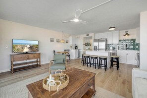 A modern living room with a television, a wooden coffee table, and a sofa. There is a kitchen area with white cabinets and a dining table.
