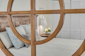 A bedroom with a wooden headboard, a bed with white bedding, a nightstand with a lamp and flowers, and white louvered closets in the background.