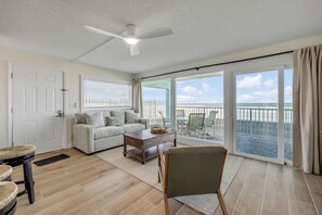 Ein geräumiges Wohnzimmer mit Sofa, Couchtisch und Sessel. Ein Deckenventilator ist sichtbar, und ein großes Schiebeglasfenster führt auf einen Balkon mit Meerblick.