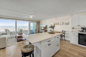 A modern kitchen with white cabinets, a dining area with a white table and chairs, and a view of the ocean through large windows.