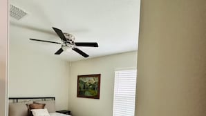 Une chambre à coucher avec un lit, un ventilateur de plafond, un tableau et une fenêtre avec des stores.