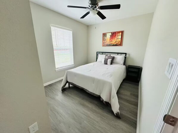 Une chambre à coucher avec un lit, un ventilateur de plafond, une fenêtre avec des stores et un tableau au mur.