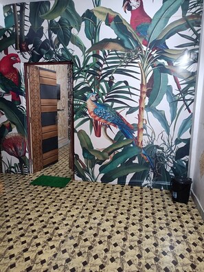 A room with a patterned floor, a door with a geometric design, and a wall mural featuring a parrot and tropical plants. - Shri Premanand Ji Niwas Sthan (Vrindavan)