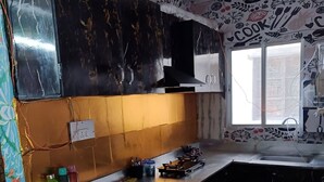 A kitchen with a patterned backsplash, a countertop with various items, and a window with a view. - Shri Premanand Ji Niwas Sthan (Vrindavan)