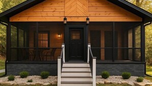 A modern log cabin with a black front door, a set of stairs leading up to the entrance, and a large windowed porch area.