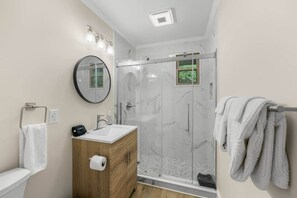 A modern bathroom with a glass-enclosed shower, a wooden vanity, a round mirror, and a towel rack. - Hill Top Pet-Friendly Cabin w/Hot Tub / Fire Pit (Pigeon Forge)