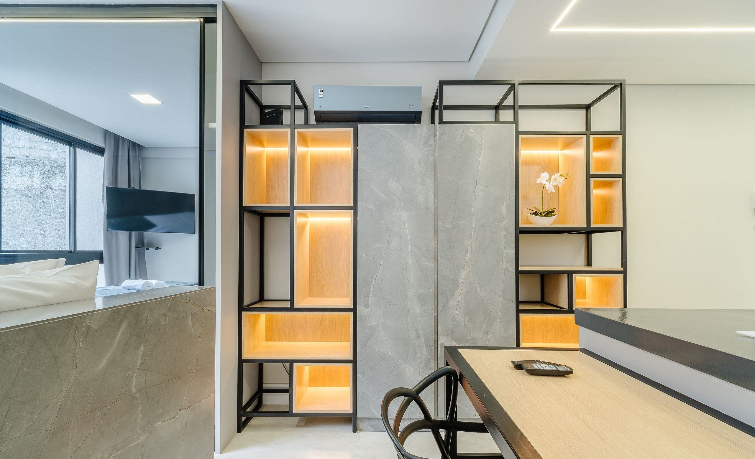 Una habitació d'hotel moderna amb un llit gran, una televisió penjada a la paret, un escriptori amb una cadira i prestatges integrats amb compartiments il·luminats.