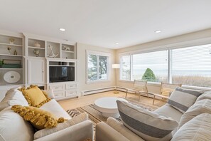A modern living room with a sofa, coffee table, and a TV. There are shelves with decorative items and a large window with blinds. - Surf Spry - Beach House in Saco Bay (Saco)