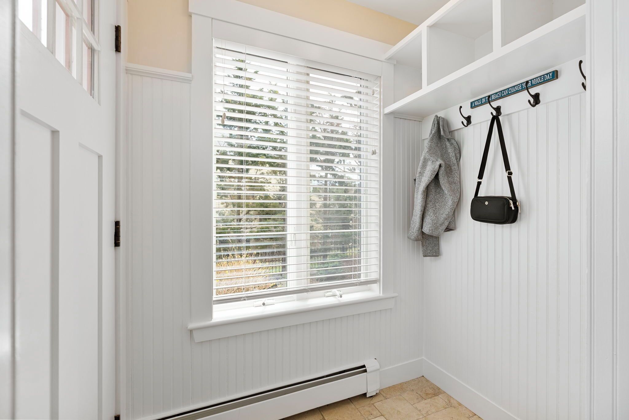 A hallway with a white door, a window with blinds, a radiator, and a coat rack with a bag and a jacket.
