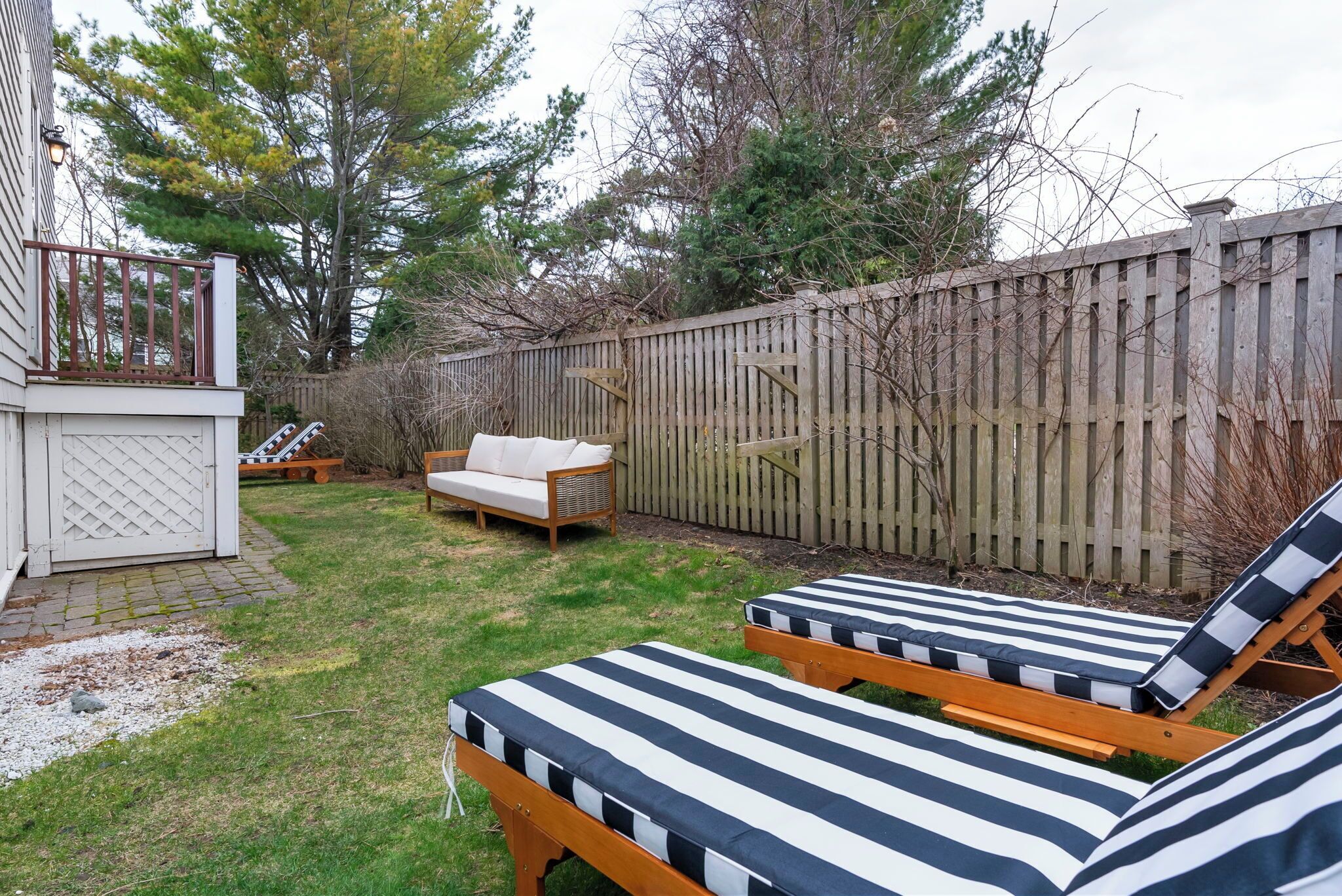 A backyard with two striped lounge chairs, a wooden deck, a white outdoor seating area, and a wooden fence.