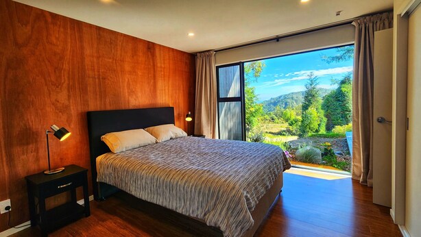 Een slaapkamer met een groot bed, houten wanden, een nachtkastje, een lamp en een glazen schuifdeur die uitkomt op een tuin.
