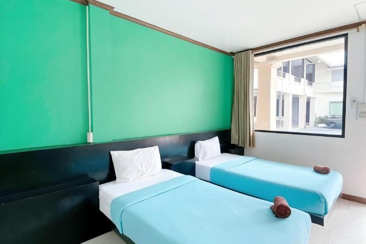 A hotel room with two beds, a green wall, a window with a view of buildings, and a curtain.