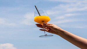 Una mano sosteniendo un vaso con pajita, que contiene un líquido amarillo, sobre un cielo despejado.