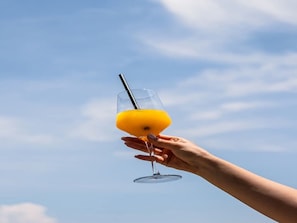 A hand holding a glass with a straw, containing a yellow liquid, against a clear sky.