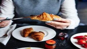 Una persona sosteniendo un croissant con un cuchillo, dos croissants sobre un plato y un frasco de mermelada sobre la mesa.