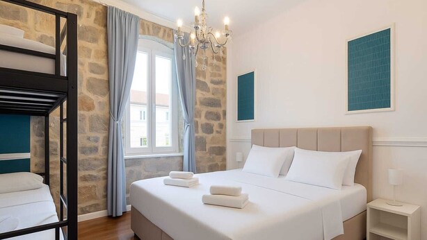 Un dormitorio con una cama nido, una cama grande con ropa de cama blanca, un candelabro, una pared de piedra y una ventana con cortinas.