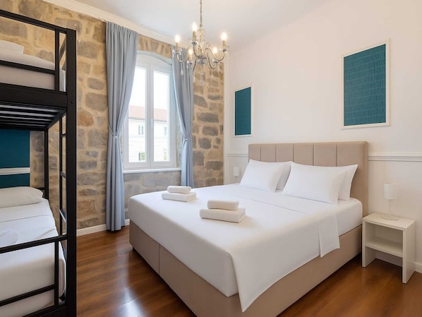 A bedroom with a bunk bed, a large bed with white linens, a chandelier, a stone wall, and a window with curtains.