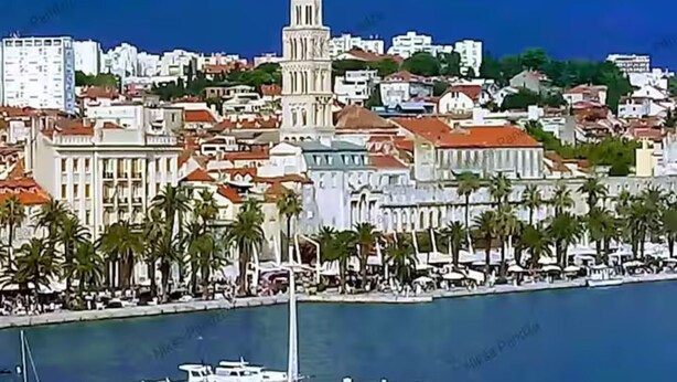 A coastal cityscape with a prominent tower, a marina with boats, and a promenade lined with palm trees.