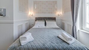 Una cama bien tendida con cabecera acolchada, dos apliques de pared y una ventana con cortinas.
